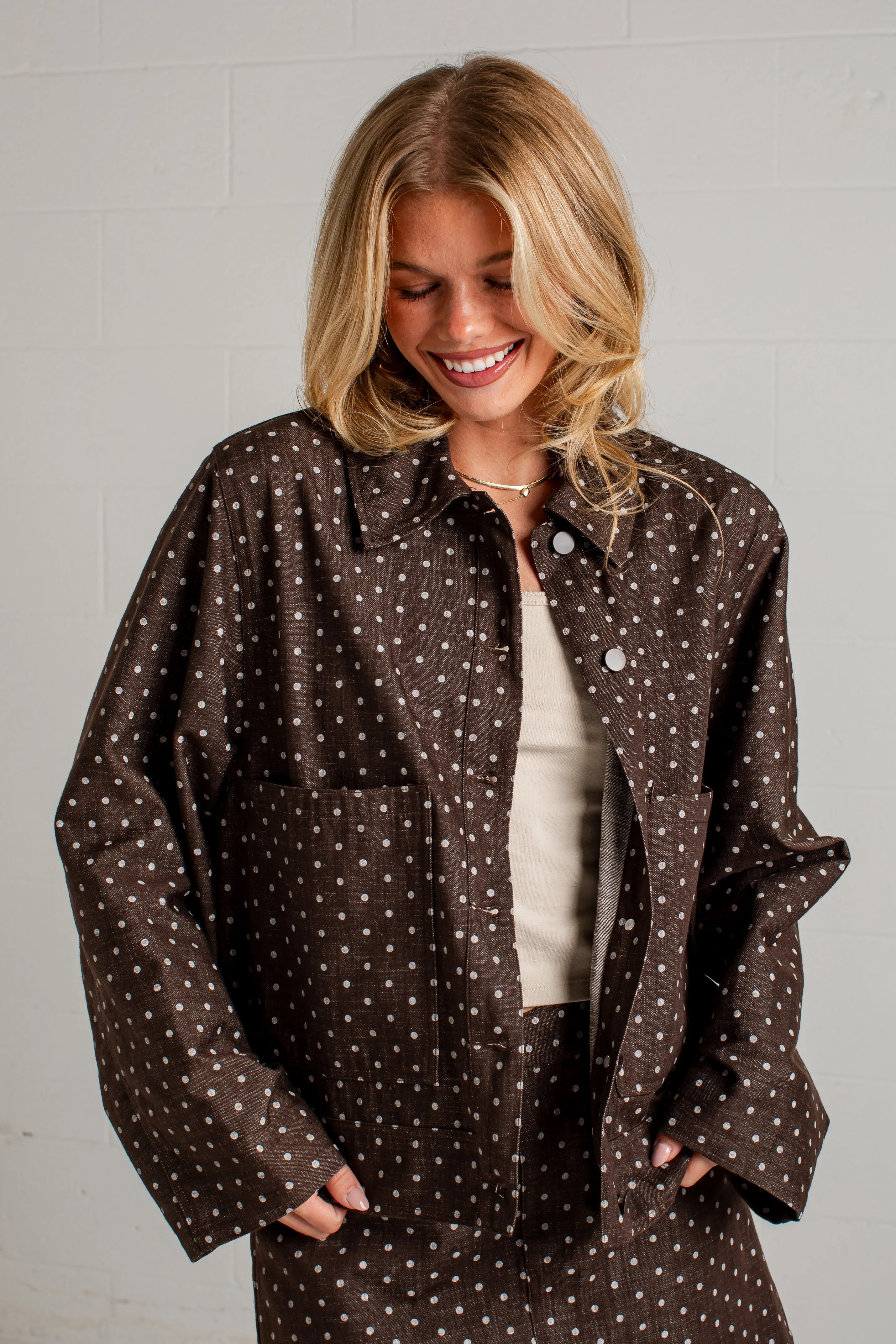 Wanita mengenakan brown polka dot jacket dari PAKAR6WIN di depan dinding batu bata putih